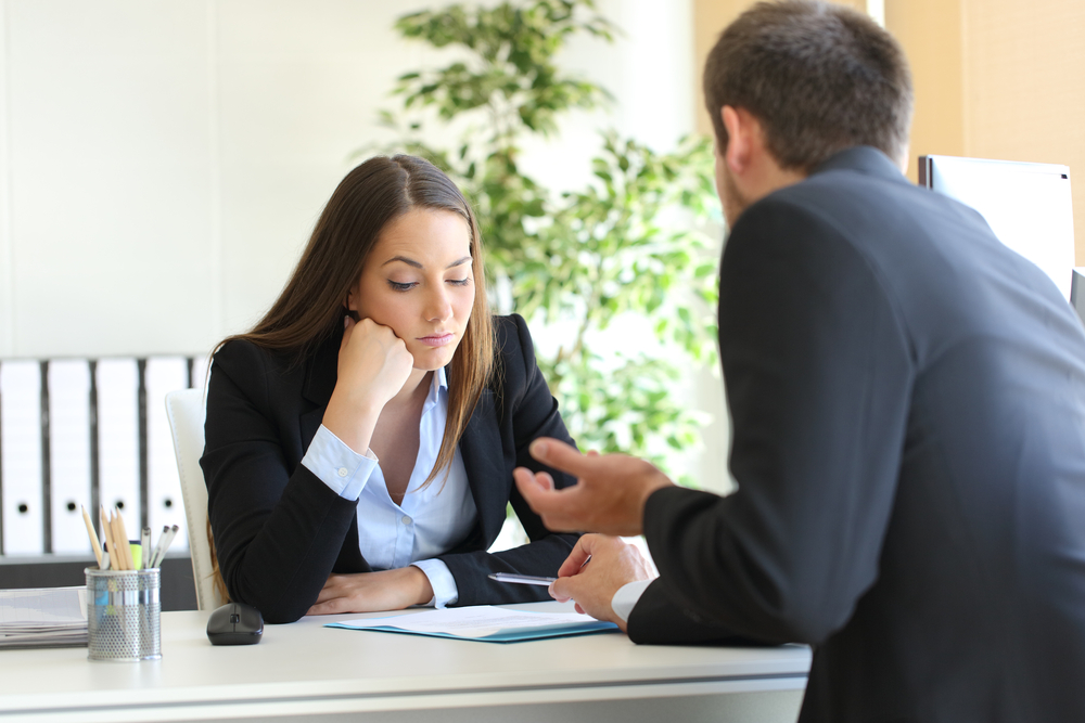 Nachdenkliche Geschäftsfrau hört einem Mann zu, beide in formeller Kleidung, am Tisch mit Dokumenten in einem hellen Büro
