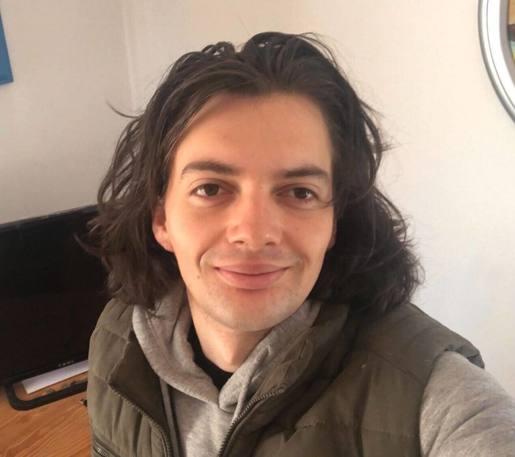 Selfie eines lächelnden Mannes mit mittellangen Haaren und einer grauen Jacke in einem Raum